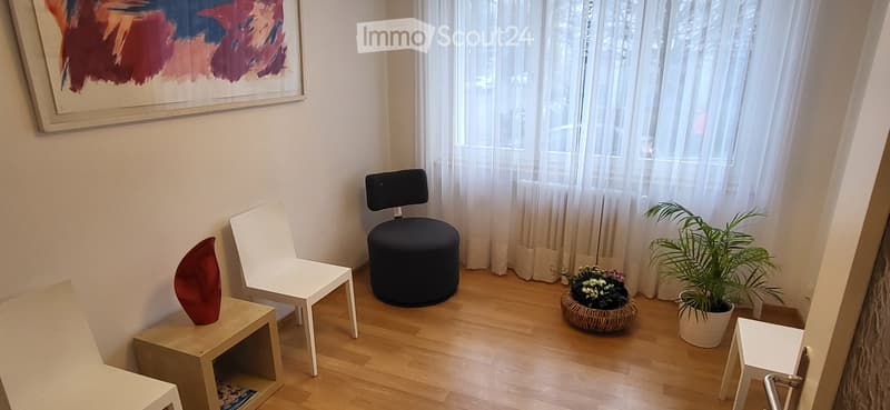Modern living room with light wooden flooring, white chairs, a black stool, small tables, a potted plant, and a decorative vase.