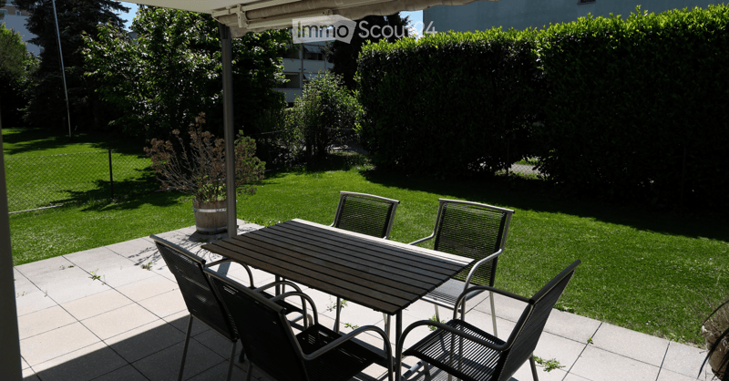 Outdoor patio with a wooden table and four black metal chairs, surrounded by a well-maintained lawn and greenery.