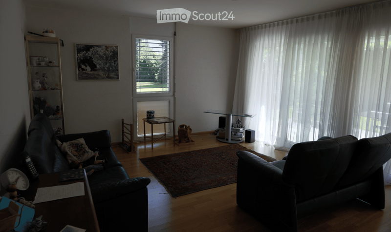 Living room with wooden flooring, large windows with white curtains, a dark leather sofa, a glass coffee table, a shelf with decorative items, and a patterned rug.