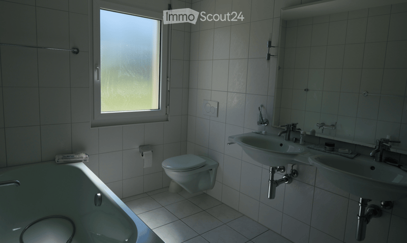 Modern bathroom with white tiles, a bathtub, toilet, double sink, mirror, and a window providing natural light.