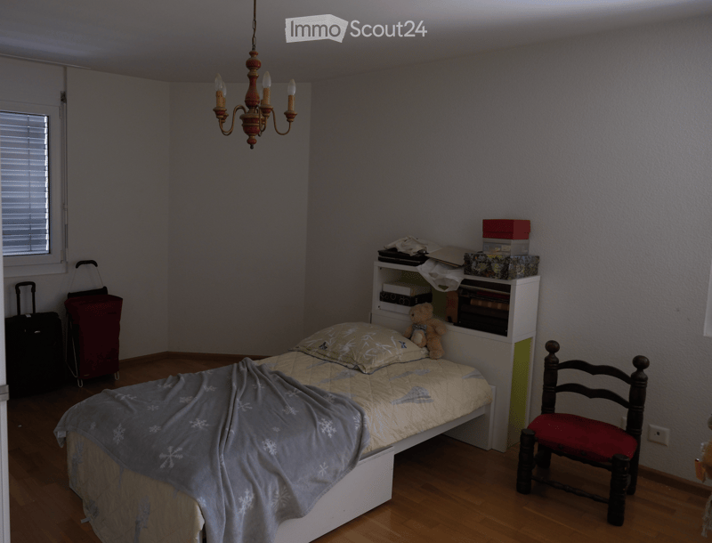 Bedroom with a single bed, a wooden chair, a shelf with books and boxes, and a teddy bear on the shelf. The bed has a patterned quilt and a blue blanket. There are two suitcases near the window.