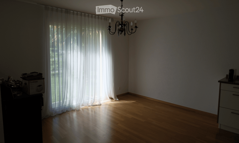 Empty room with wooden flooring, large window with white curtains, and a chandelier hanging from the ceiling. No furniture or furnishings are present.