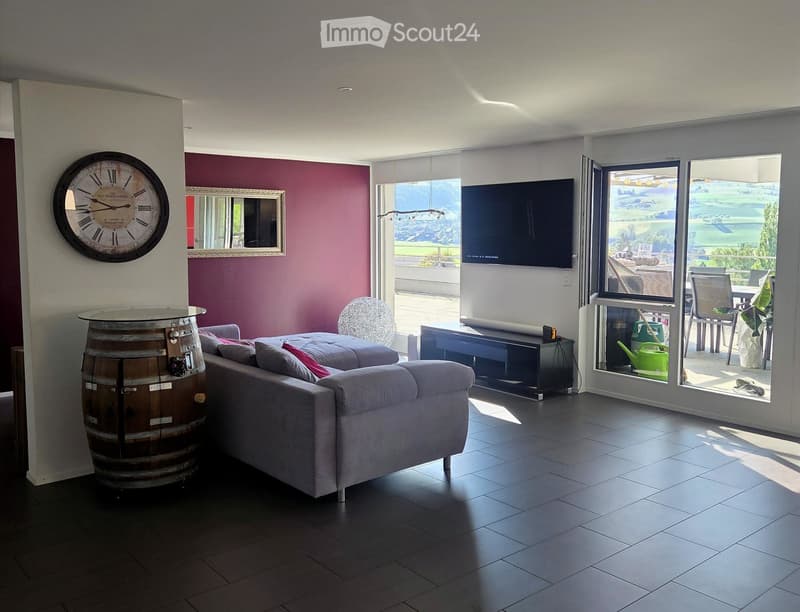 Modern living room with grey sofa, coffee table, wall clock, television, and large windows offering a scenic view.
