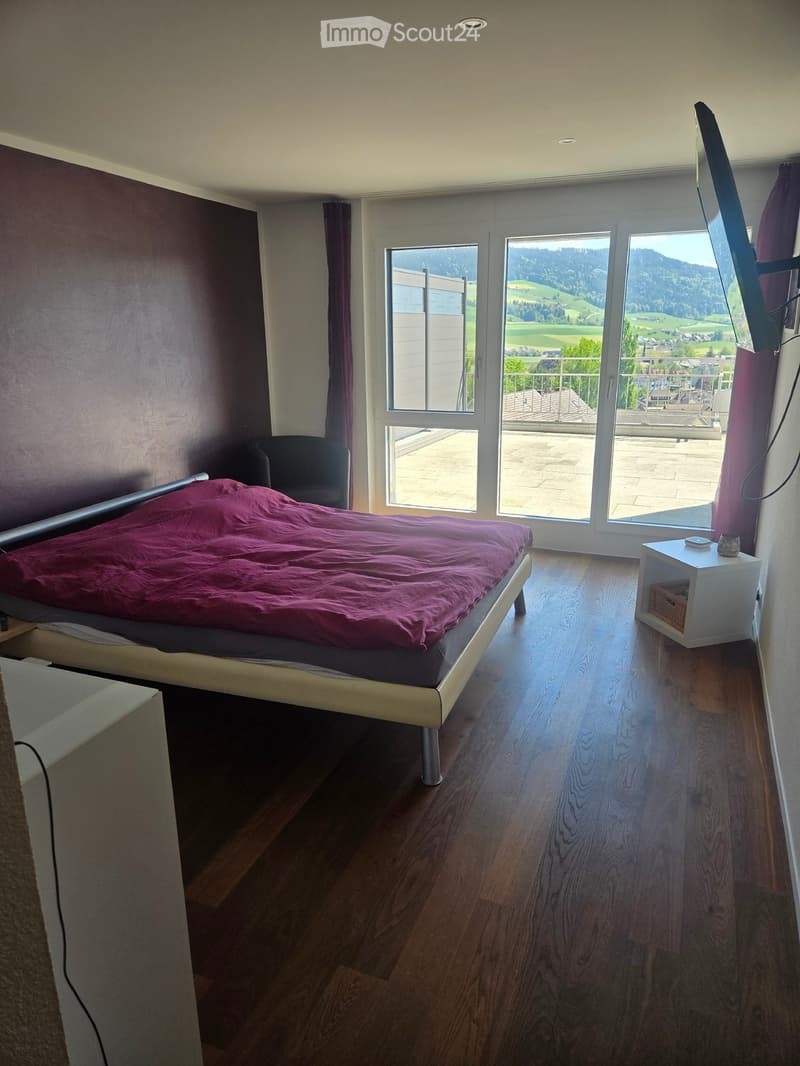 Modern bedroom with wooden flooring, large windows, and a balcony view. Features a bed with a purple duvet, a nightstand, and a chair.