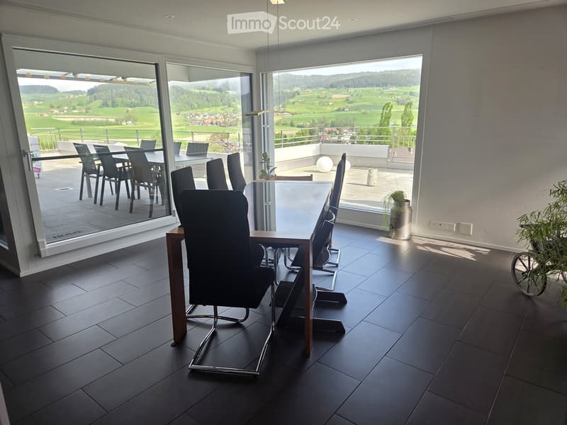 Modern dining area with dark tiled floor, large glass windows offering scenic countryside view, and a table with chairs.