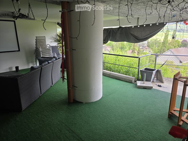 Balcony with green artificial turf, white columns, a sofa, chairs, a small table, and a screen mounted on the wall. The balcony has a metal railing and offers a view of greenery and houses.