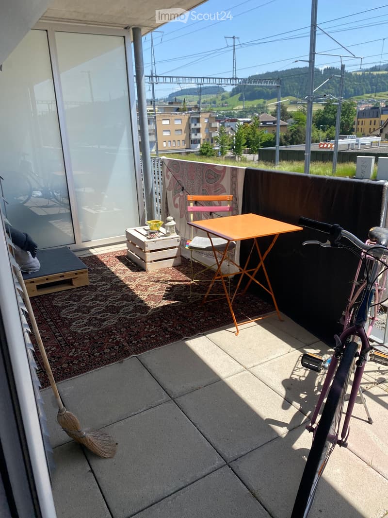 Balcony with a view, featuring a small orange table, two chairs, a bicycle, a rug, a wooden stool, and a lamp.