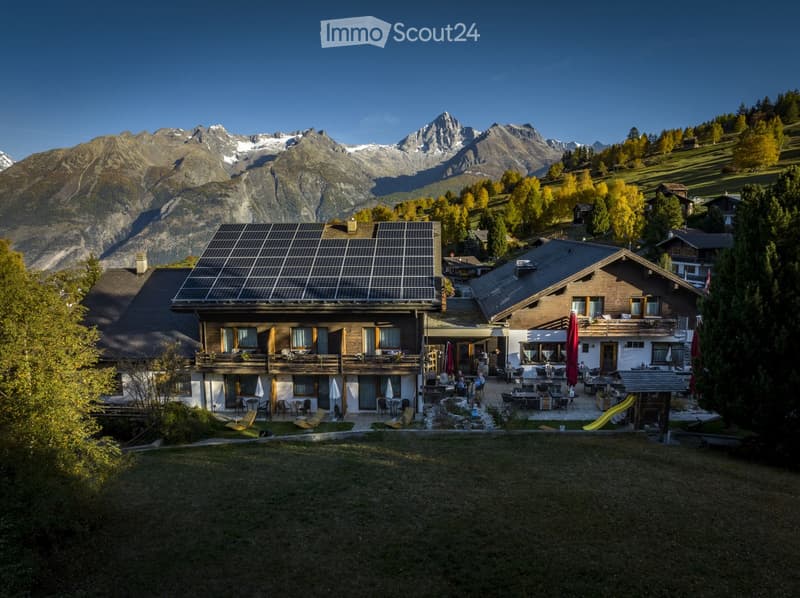 A large, modern house with a balcony and solar panels on the roof, surrounded by a well-maintained lawn and outdoor seating area, with mountain views in the background.