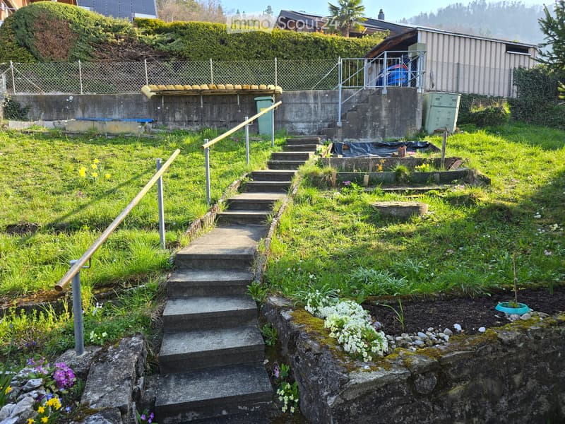 A sloped garden area with concrete steps, metal railings, and various plants and flowers. The garden is bordered by a concrete retaining wall and a fence, with a small shed visible in the background.