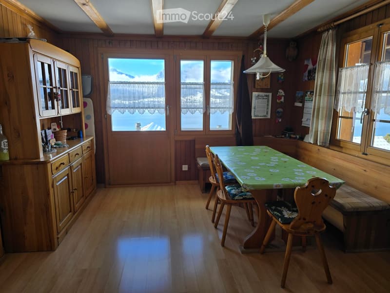 Cozy kitchen with wooden walls and floors, large windows offering a scenic mountain view, a dining table with chairs, and wooden cabinets.