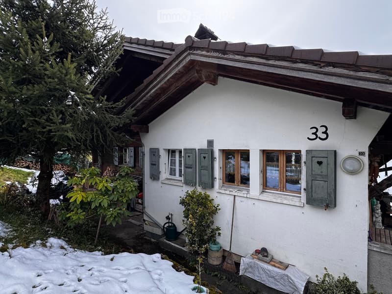 A two-story white house with wooden shutters and a sloped roof, featuring the number "33" on the facade. The house is surrounded by greenery and snow-covered ground.