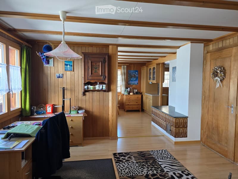 A well-lit hallway with wooden walls and floors, featuring a desk with a chair, a hanging lamp, a cabinet, and a kitchen area visible in the background.