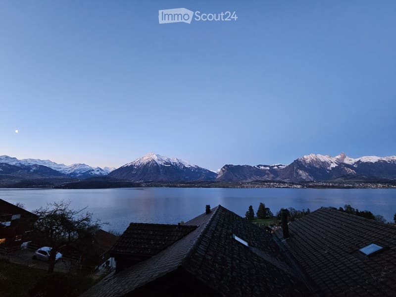 A view of a house with a stone roof and chimney, overlooking a lake and snow-capped mountains in the background.