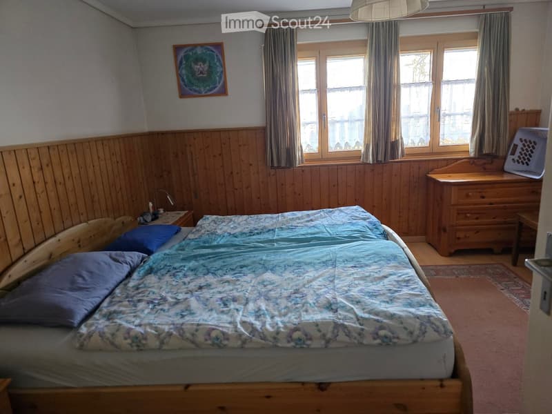 Cozy bedroom with wooden walls, a large bed with blue bedding, a nightstand with a lamp, a wooden dresser, and a framed picture on the wall.