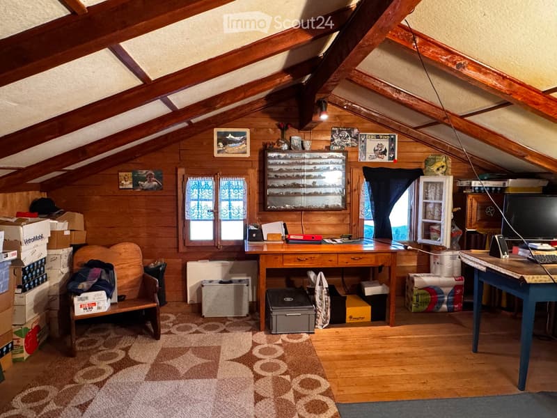Cozy attic room with wooden beams, wooden walls, and a slanted ceiling. Features a wooden desk, a chair, a small bench, a table with a screen, a glass-fronted cupboard, and several boxes and items stacked around.