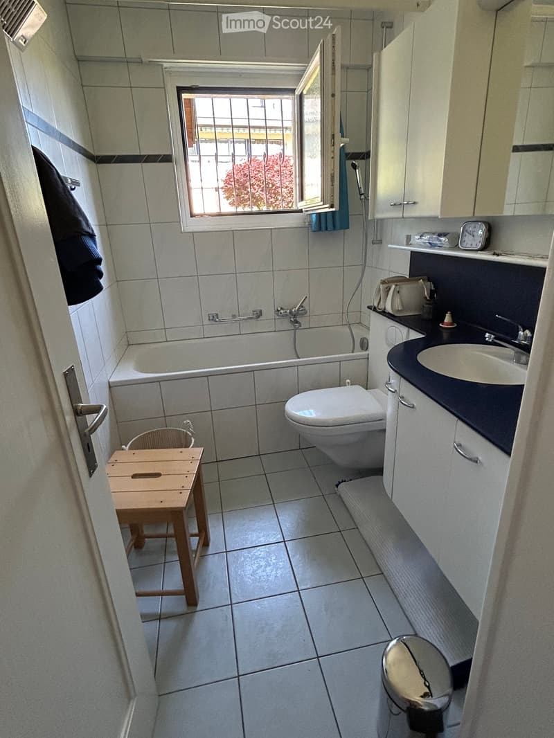 Bathroom with white tiles, a toilet, a sink with a cabinet, a bathtub, a small wooden stool, and a window with a view of a tree.