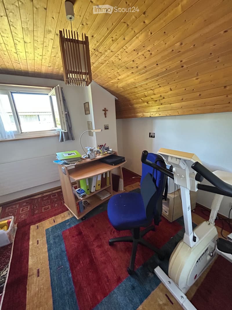A cozy attic room with wooden beams, a large window, a desk with a chair, an exercise bike, and a colorful rug.