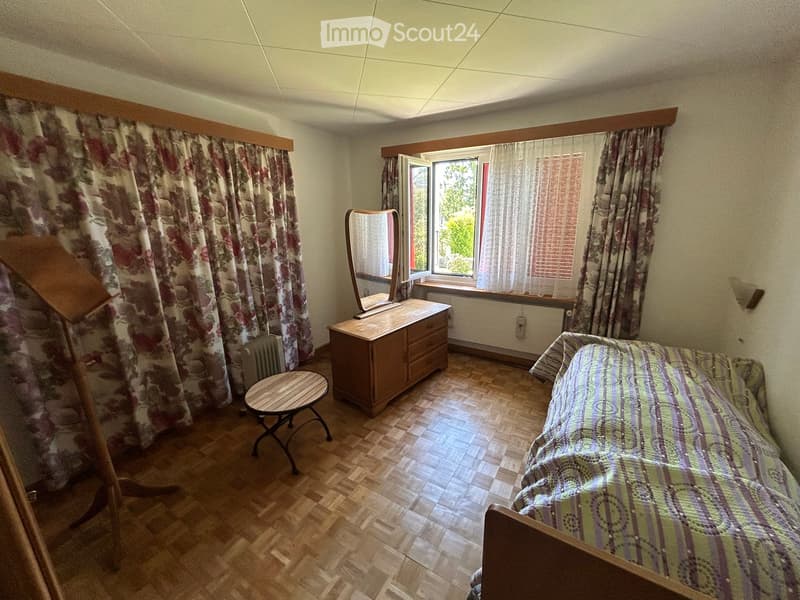Bedroom with wooden flooring, a bed with a patterned bedspread, a wooden dresser with a mirror, a small round table, and floral curtains on the windows.