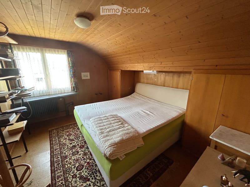 Cozy bedroom with wooden walls and ceiling, featuring a single bed with a green base, a folded blanket, a small table with toys, a chair, and a shelf with books and a computer.