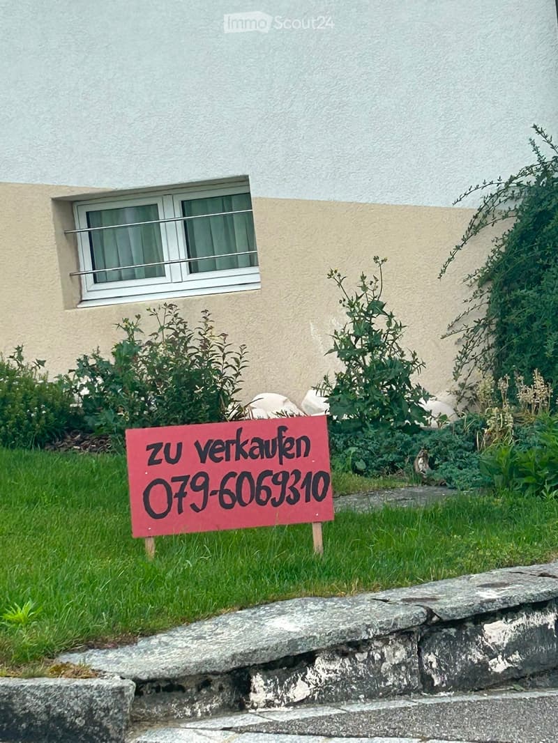 Exterior view of a two-story house with a window, small garden, and a "zu verkaufen" sign indicating it is for sale.