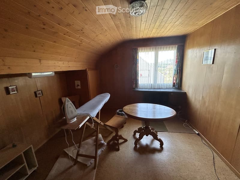 Small room with wooden walls and ceiling, featuring a round wooden table, an ironing board with an iron, and a small stool.