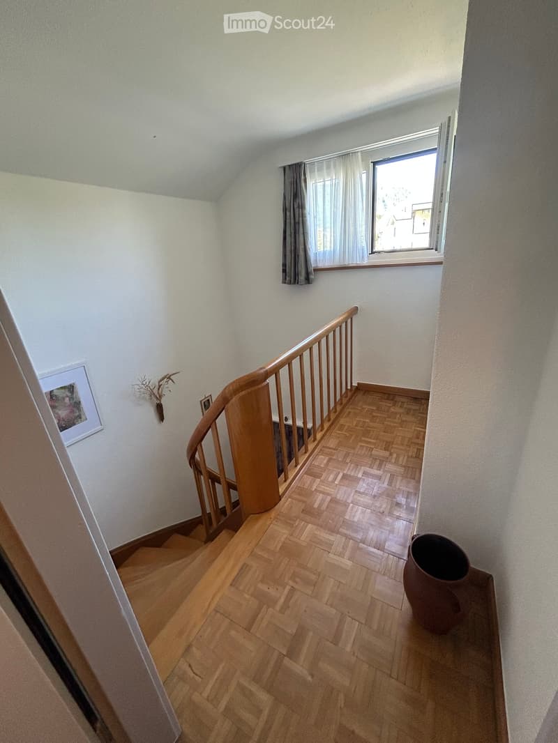 Spacious hallway with wooden herringbone flooring, curved staircase with wooden railing, large window with curtains, and a decorative vase on a small table.