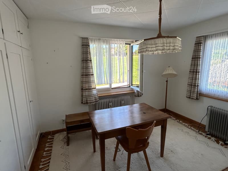 A furnished room with a wooden table, chairs, a floor lamp, and radiators. The room has large windows with curtains and a patterned rug on the floor.