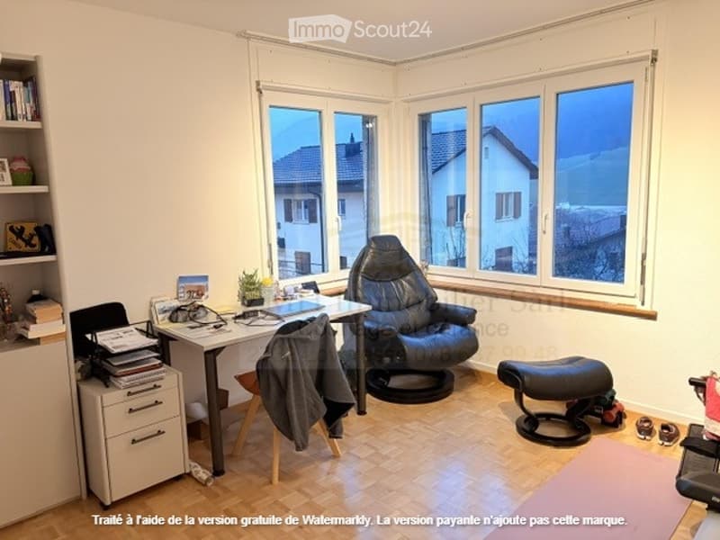 A room with wooden floor, white walls, large windows, a desk, black leather chair, footstool, books, a cabinet, storage boxes, clothes, and other items.