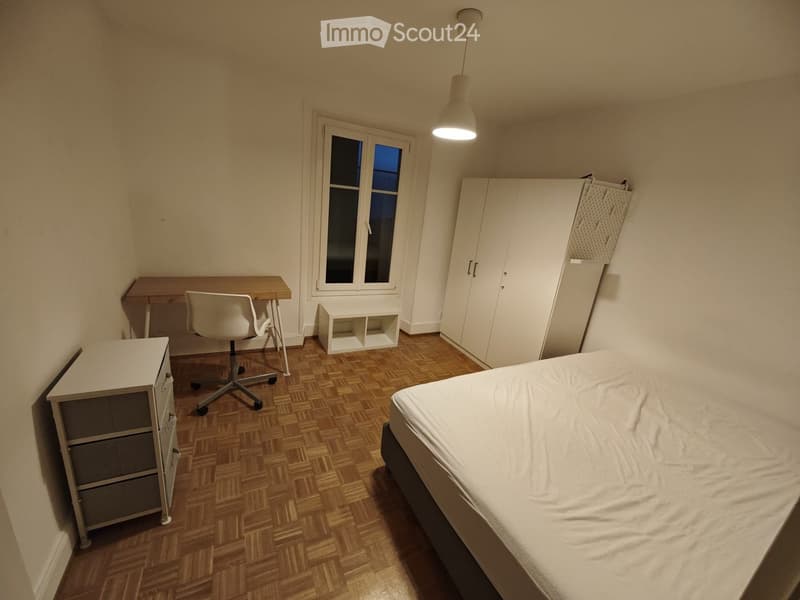 A well-lit bedroom with a wooden floor, a bed, a desk with a chair, a cabinet, and a small shelf near the window.