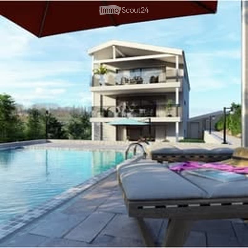 Modern two-story building with balconies, surrounded by greenery, with a swimming pool and lounge chairs in the foreground.