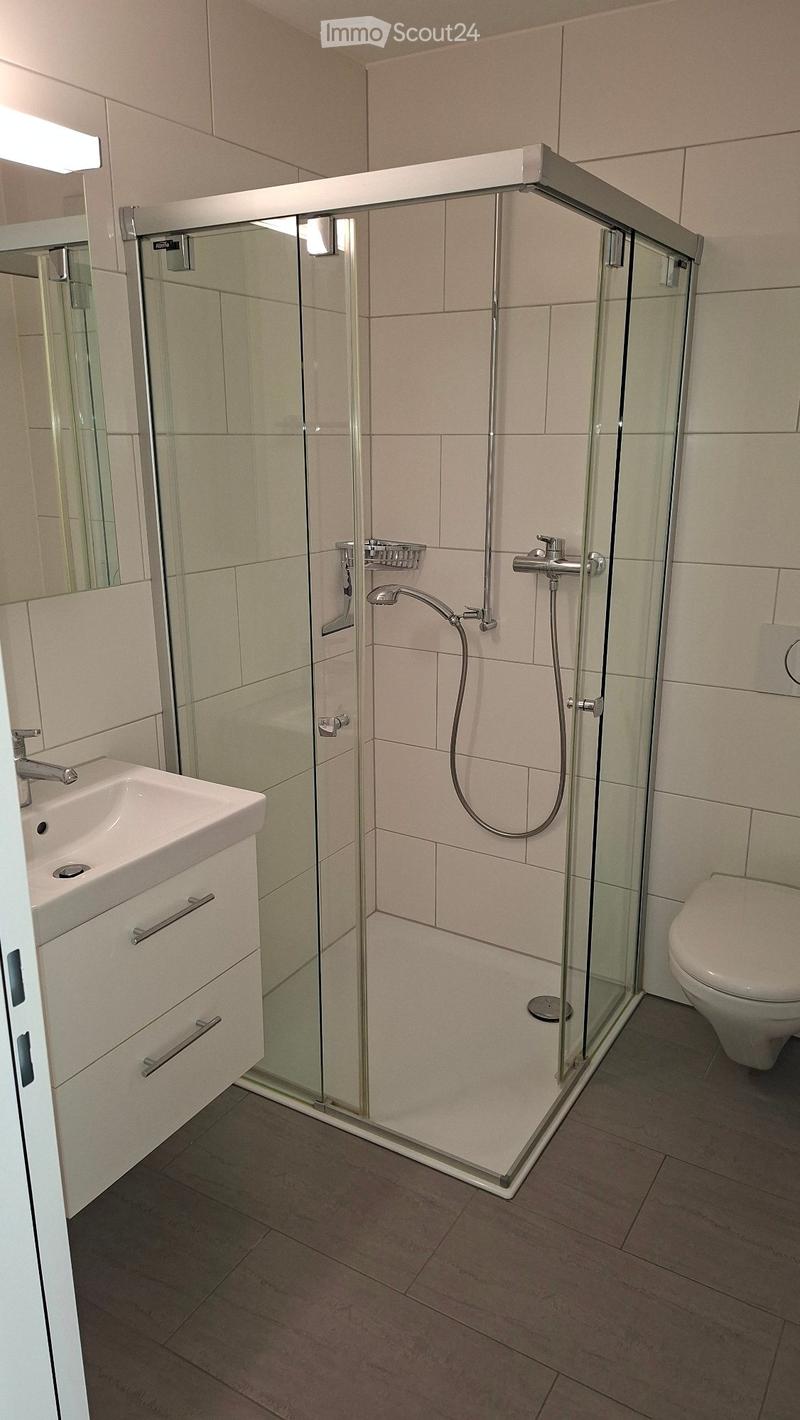 Modern bathroom with a glass-enclosed shower, a white sink with a cabinet, and a toilet. The walls are tiled in light beige, and the floor is covered with dark tiles.