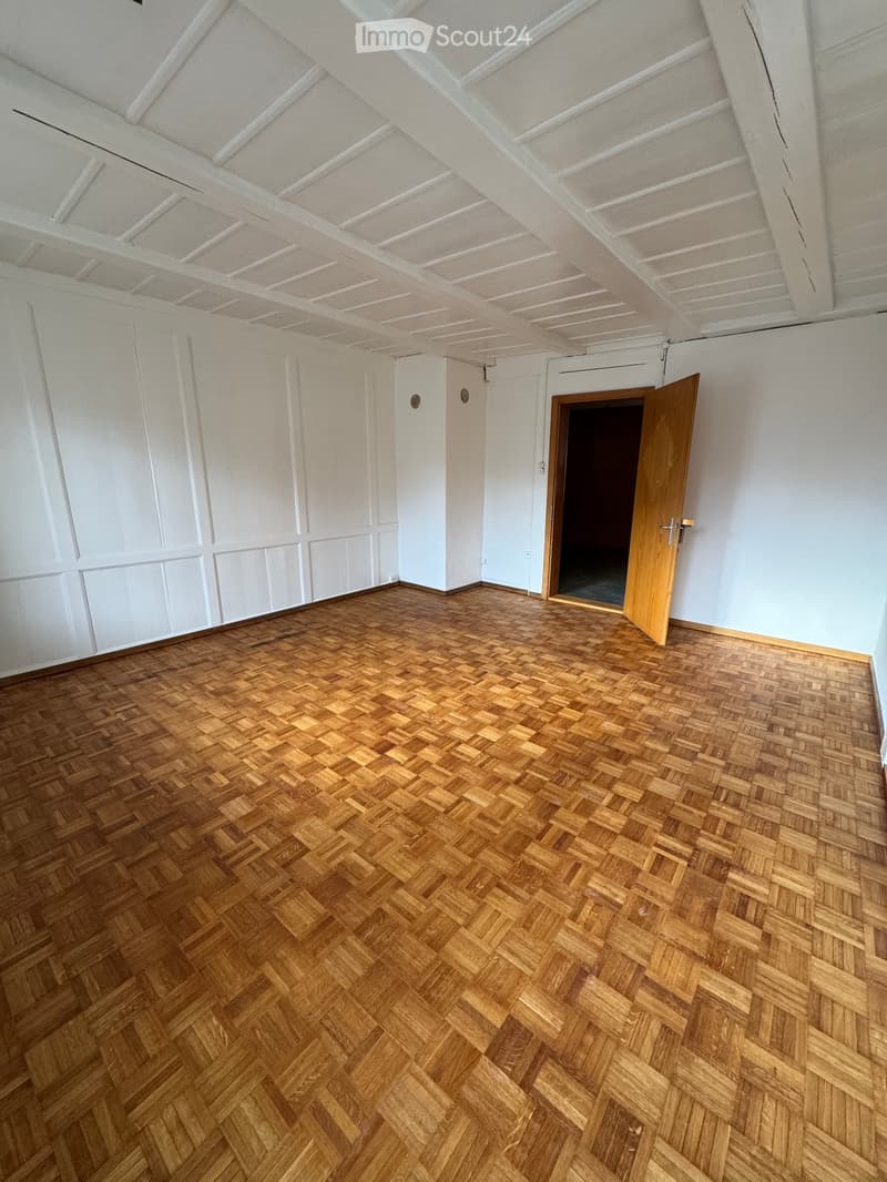 Empty room with wooden herringbone flooring, white walls, and a wooden door leading to another space.