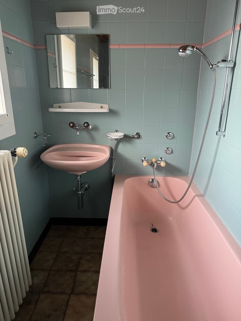 Bathroom with a pink bathtub, pink sink, mirror, and radiator. Light blue tiled walls with pink trim.