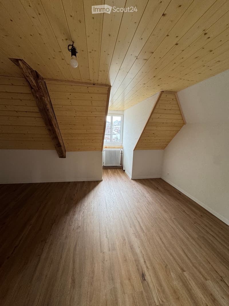 Empty attic room with wooden floors, slanted wooden ceiling, and a single window showing a view of the outside.