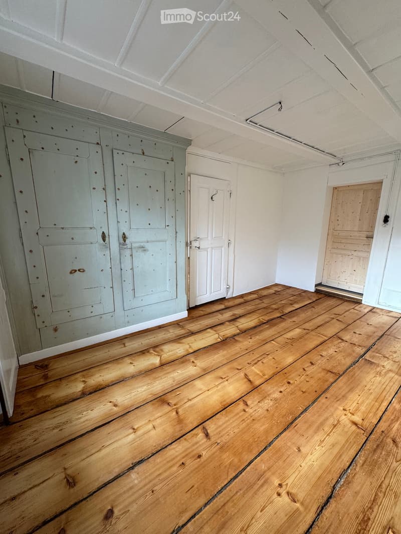 Empty room with wooden flooring, white walls, and several doors including a large wooden wardrobe and a smaller white door.