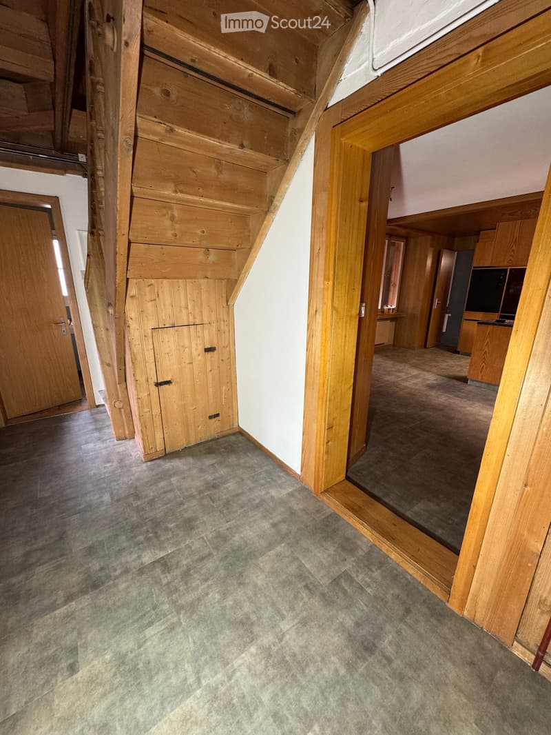 Empty hallway with wooden beams and doors, leading to a kitchen area with cabinets and appliances.