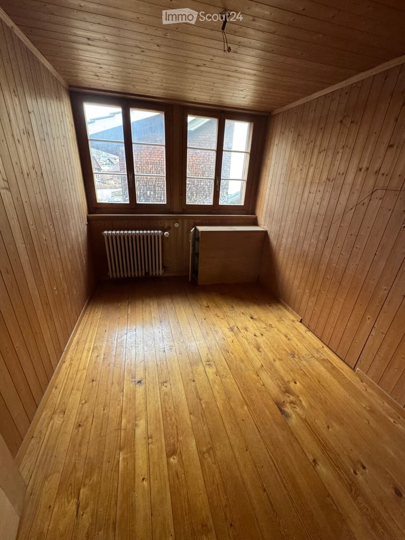 Empty room with wooden walls, wooden floor, and large windows showing a view of the outside. There is a radiator on the left side and a small cabinet on the right side.