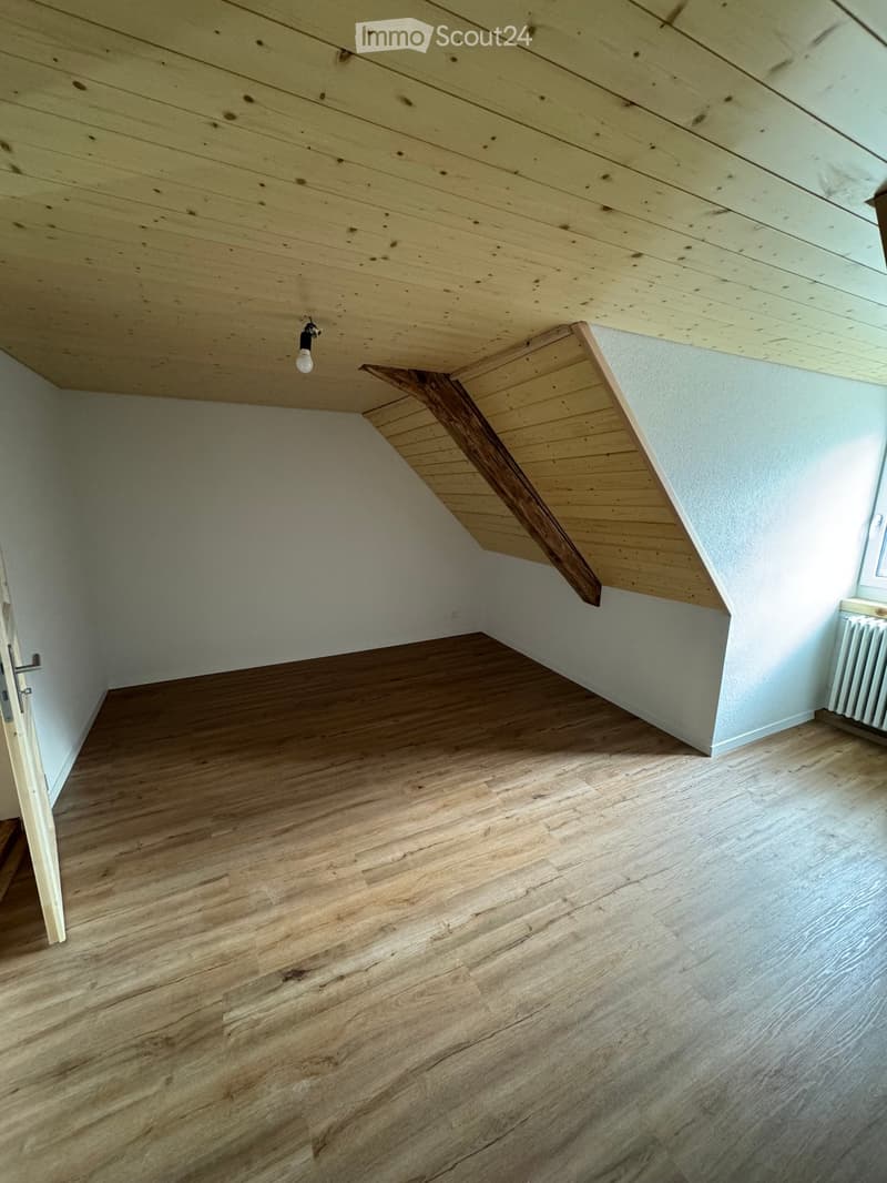 Empty attic space with wooden flooring, slanted wooden ceiling, and a single light fixture. The room has white walls and a small window on one side.