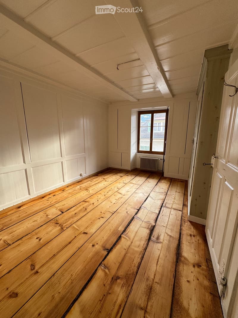 Empty room with wooden flooring, white walls, and a window showing a view of a building outside. The room has a simple, clean design with no furniture or decorations.