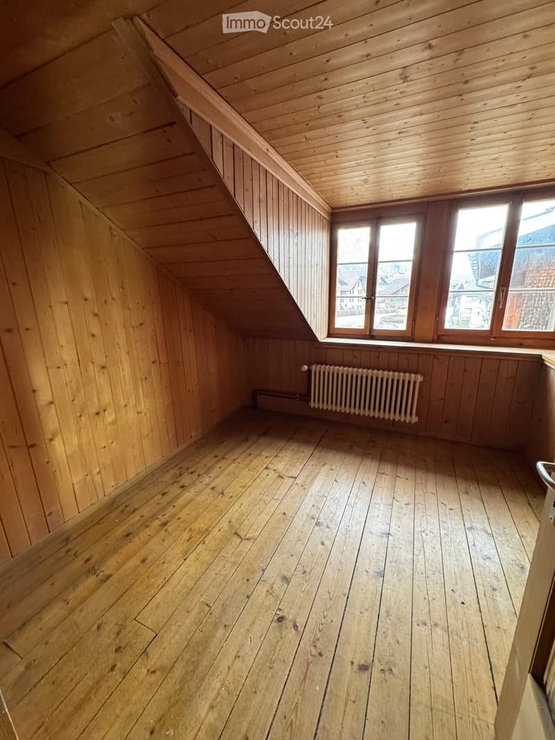 Empty attic room with wooden walls, wooden floor, and a single window with a radiator below it.