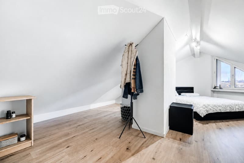 Bright attic bedroom with wooden flooring, slanted white ceilings, large windows, a bed with a patterned duvet, a nightstand, a wooden shelf, a coat rack with jackets, and a small basket on the floor.
