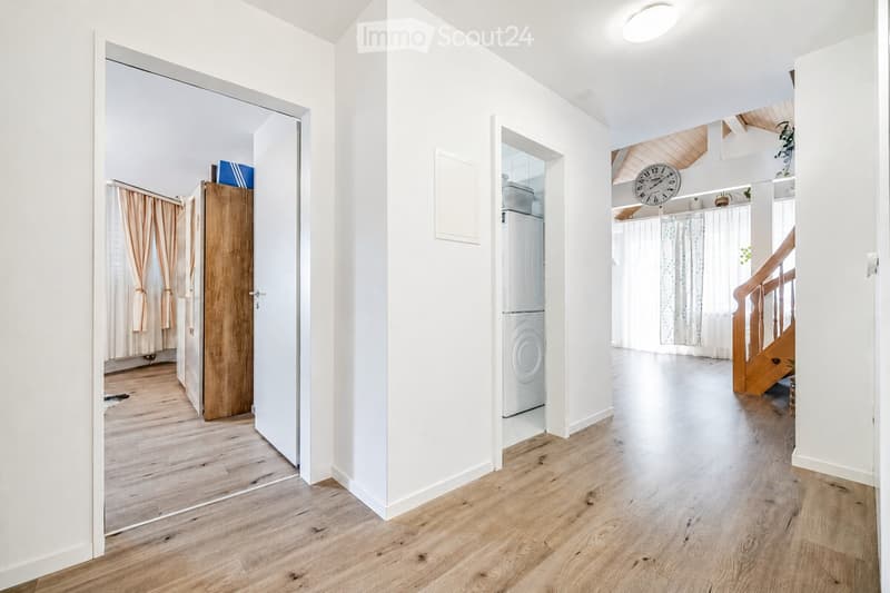 Bright hallway with wooden flooring, white walls, and open doors leading to adjacent rooms. Features a staircase with wooden railings and a clock on the wall.