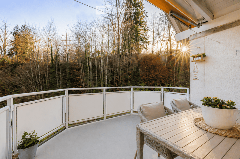 Balcony with a wooden table, wicker chairs, and potted plants, overlooking a wooded area with sunlight streaming in.