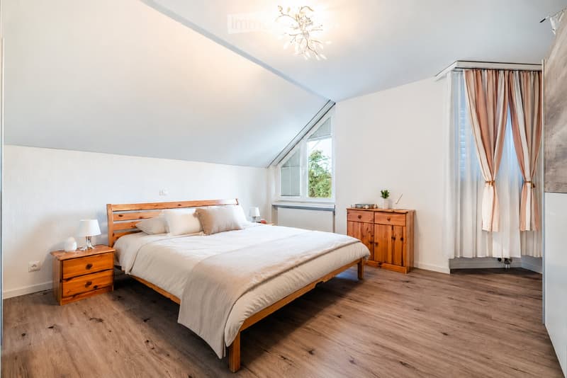 Cozy bedroom with wooden bed, nightstands, lamps, and a wooden cabinet. Light-colored bedding and curtains complement the light wood flooring and white walls.