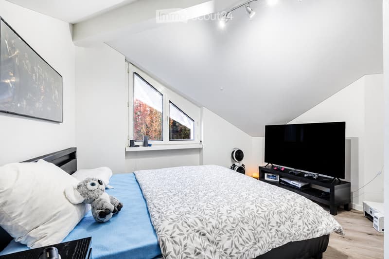 Bright, modern bedroom with a large bed, a TV stand, and a stuffed animal on the bed. The room features large windows with a view, a sleek TV, and a cozy atmosphere.