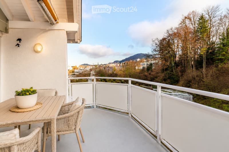 Balcony with a view of the city and surrounding landscape, featuring a small table, chairs, and a flower pot.