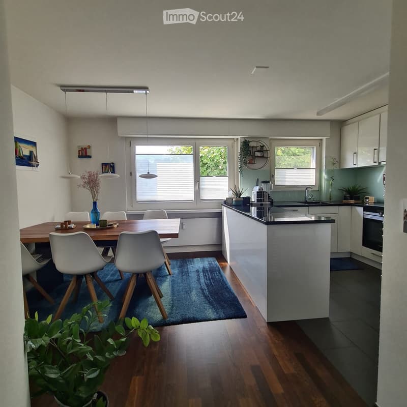 Open-plan living and dining area with a wooden dining table, white chairs, a modern kitchen island, and various appliances.