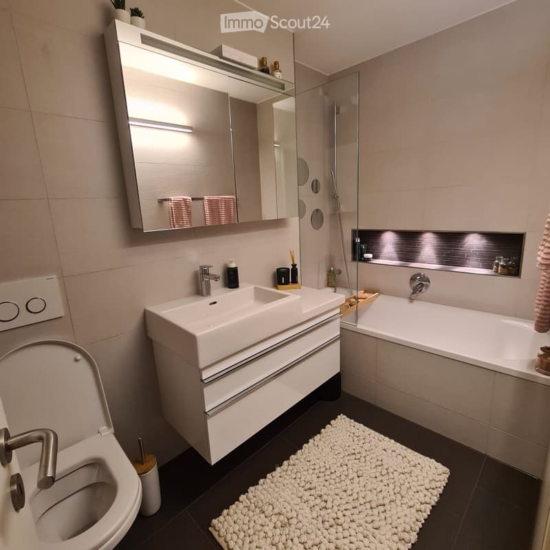 Modern bathroom with a white sink, toilet, bathtub, and a large mirror with built-in lighting. The space features beige tiles, a white rug, and various toiletries on the sink counter.