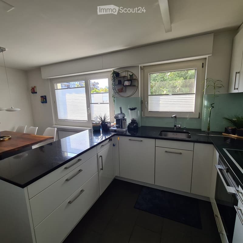 Modern kitchen with white cabinets, black countertops, and stainless steel appliances. Features a coffee maker, blender, sink, and a small dining area with chairs and a table.
