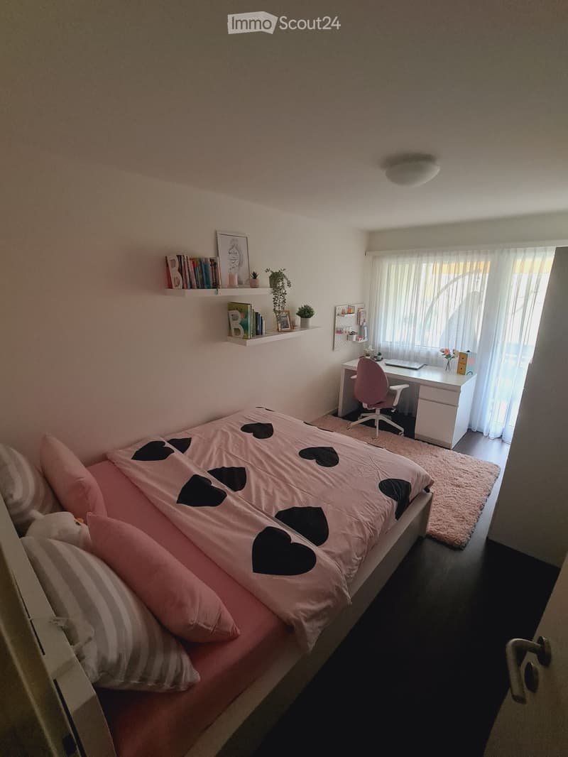 Cozy bedroom with a bed covered in pink and black heart-patterned bedding, a white desk with a chair, a shelf with books and decorative items, and a window with sheer curtains.